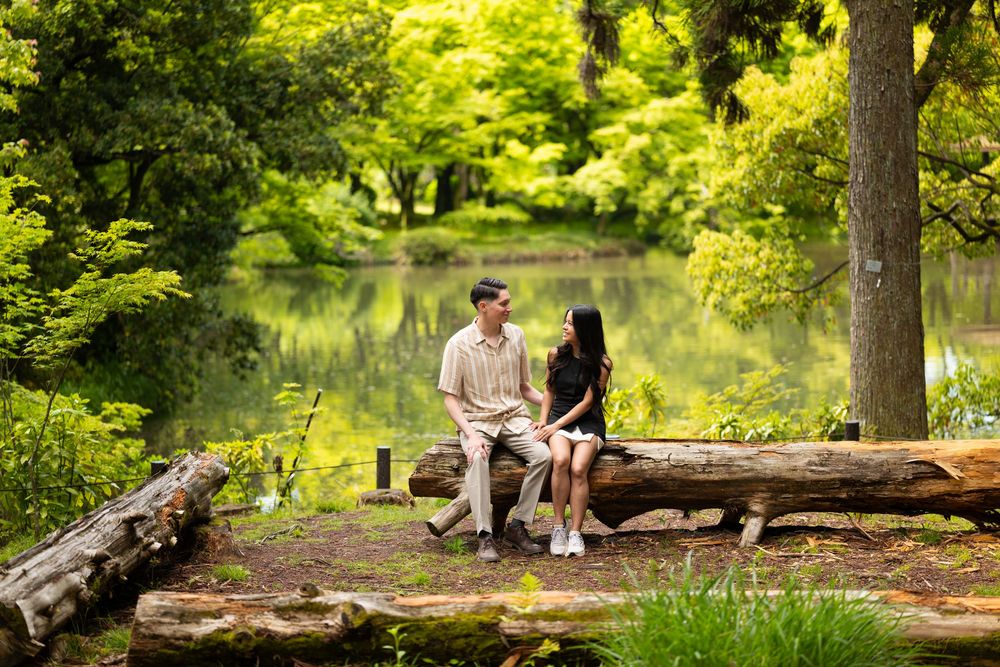 Couple sitting together on a weathered log beside a calm pond, surrounded by lush green trees in a peaceful park.