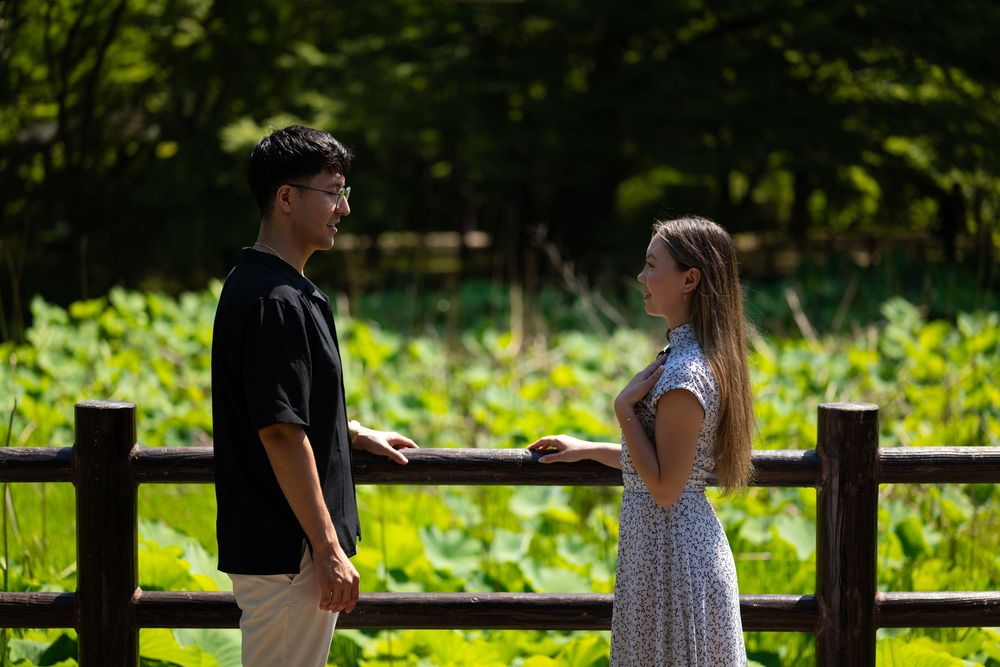 A smiling man in a black shirt and a woman in a patterned dress stand facing each other by a wooden fence with lush green plants behind.