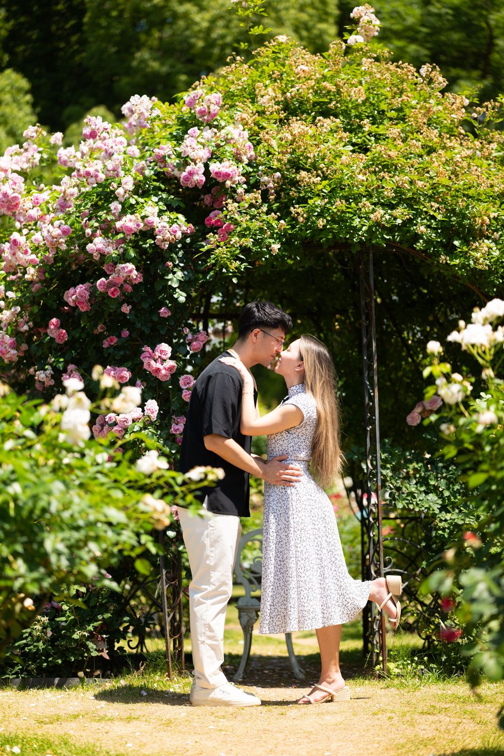 Couple sharing a kiss beneath a rose-covered garden arch, surrounded by pink and white blooms on a sunny day.