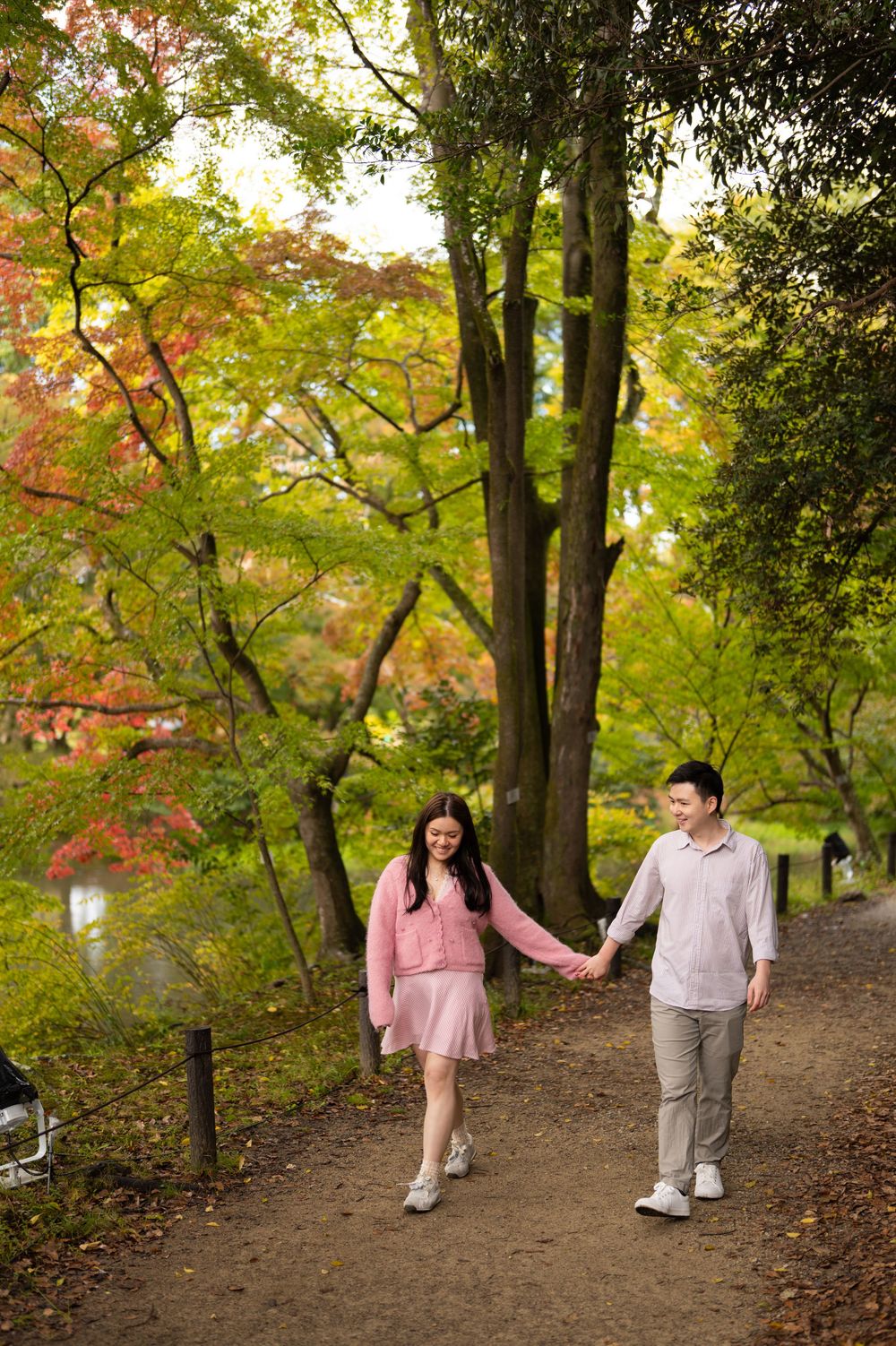 A smiling couple holds hands while walking along a dirt path surrounded by colorful autumn trees in a peaceful park.
