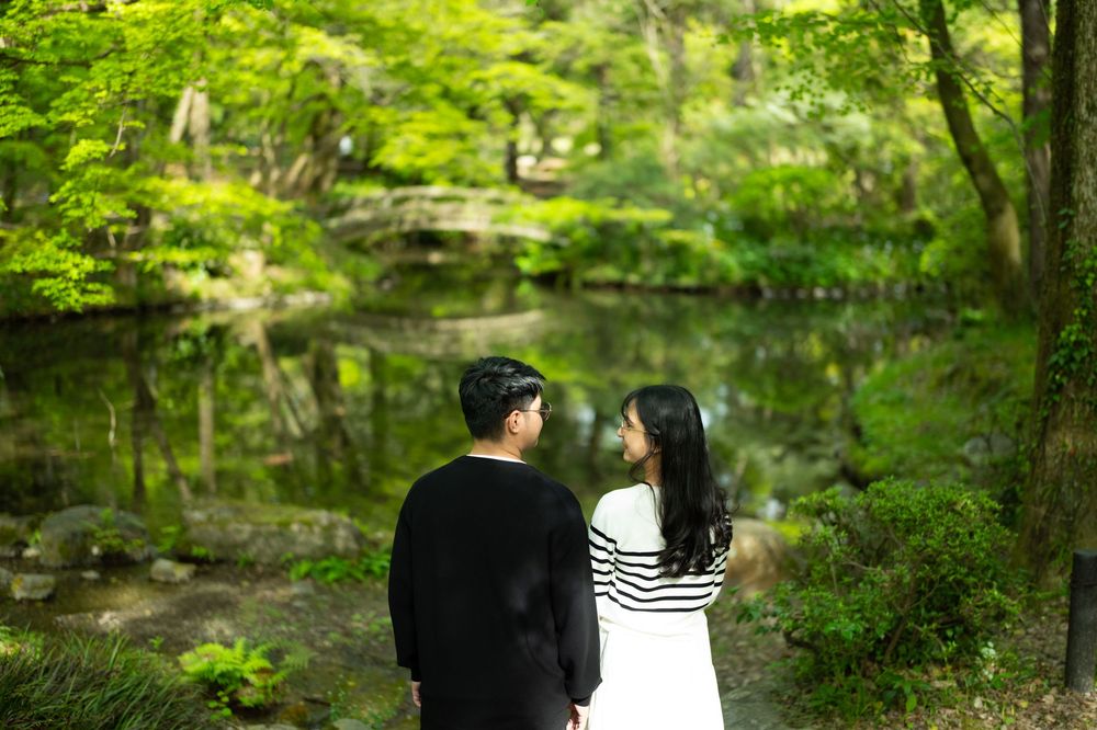 Couple in dark and white outfits stand facing each other beside a calm pond in a lush green Japanese garden.