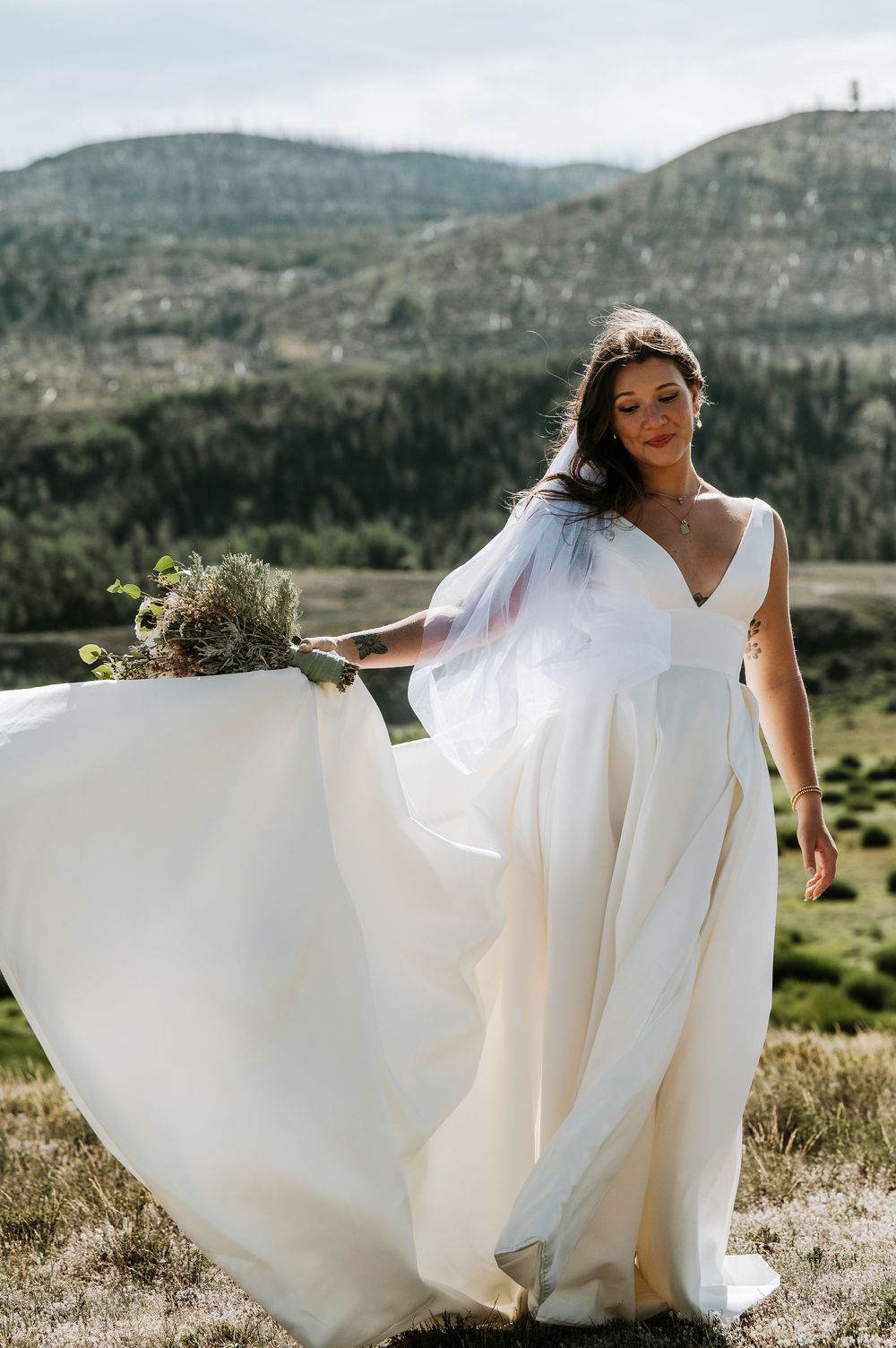 Bride in a flowing white gown and veil holds a wildflower bouquet while standing in a grassy mountain meadow with forested hills behind.