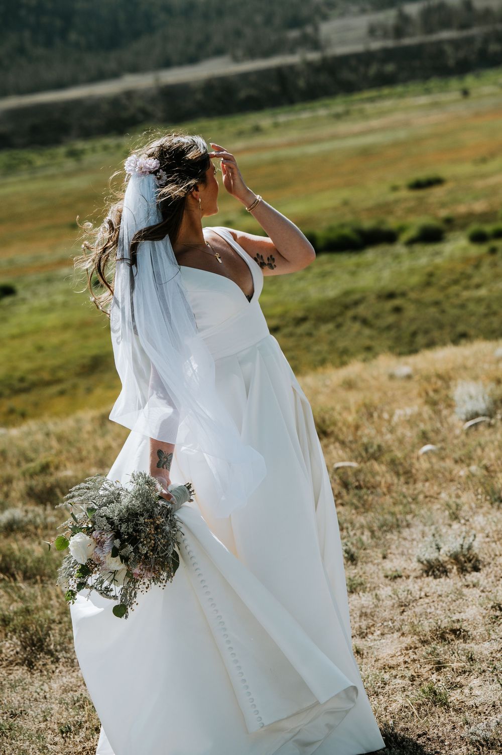 Bride in a white satin gown and flowing veil holds a wildflower bouquet while standing in a sunlit grassy mountain meadow.