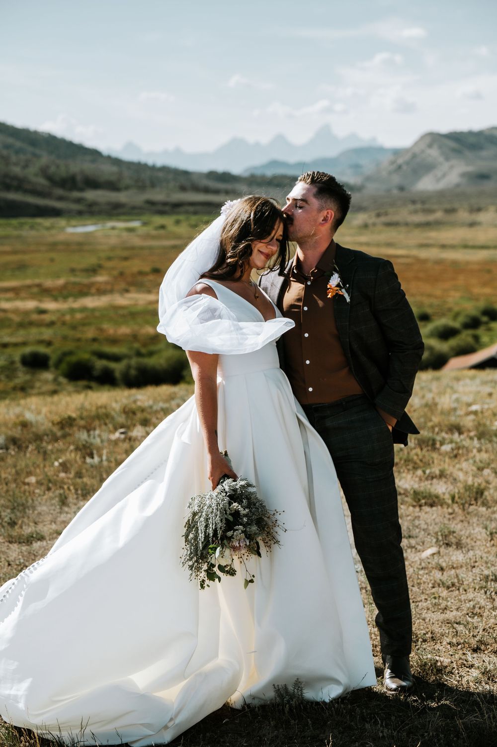 Bride in a flowing white gown holding a bouquet is kissed on the forehead by her groom in a mountain meadow elopement.