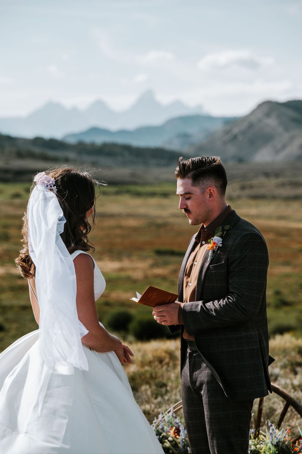 Bride in a white gown and veil faces a groom in a plaid suit reading vows from a book, with mountains behind them.