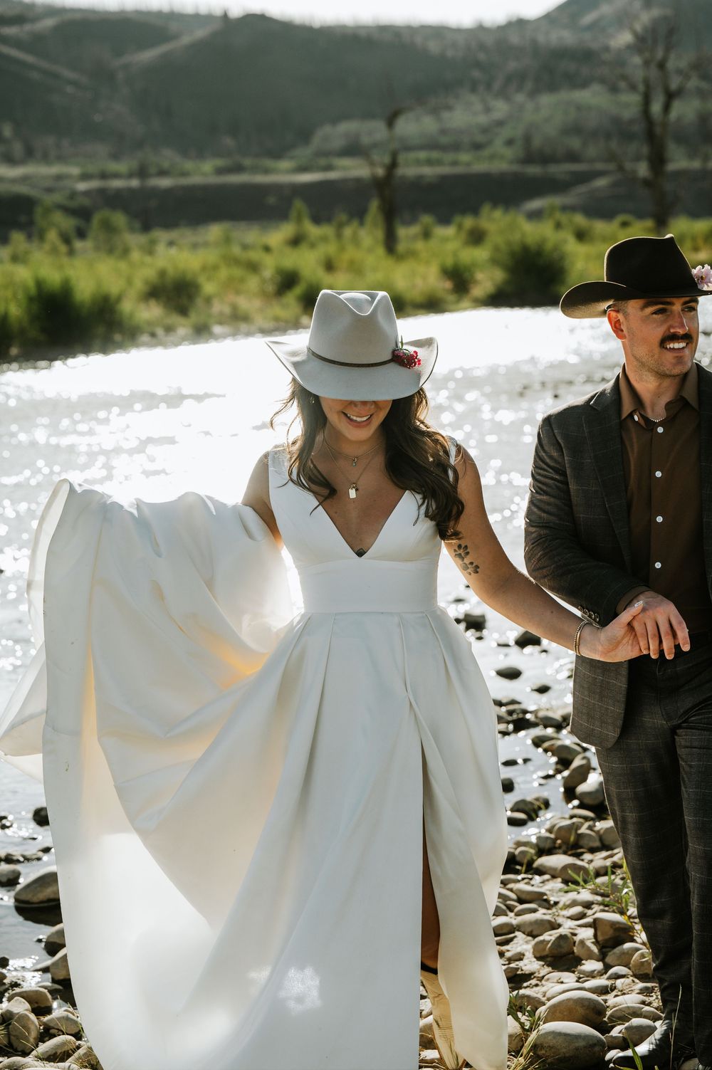 Bride in a white gown and gray felt hat walks hand in hand with a groom in a black hat along a rocky riverbank during a mountain elopement.