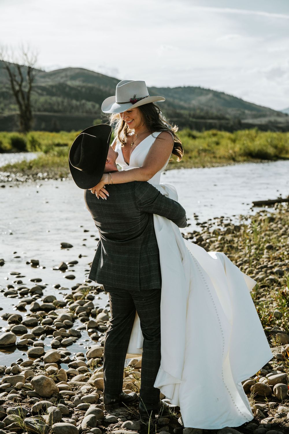 Groom in a black cowboy hat lifts his bride in a white gown and gray hat beside a rocky riverbank with mountains behind.