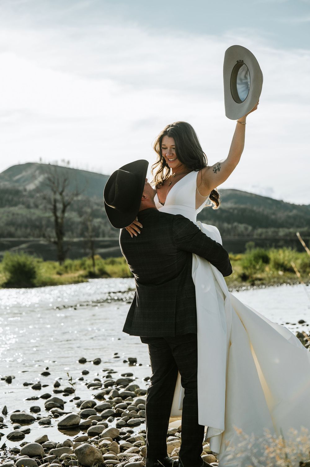 Groom in a black cowboy hat lifts his bride in a white gown as she waves a gray hat on a rocky riverbank with mountains behind.