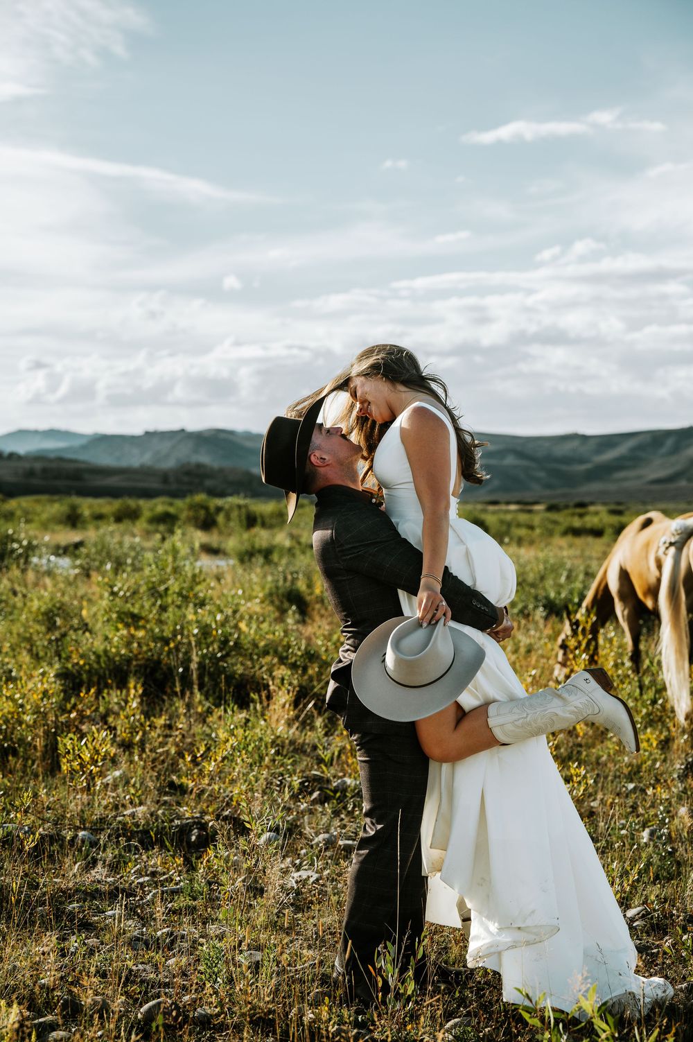 A groom in a black cowboy hat lifts and kisses his bride in a white gown and boots in a grassy field with a horse and mountains.