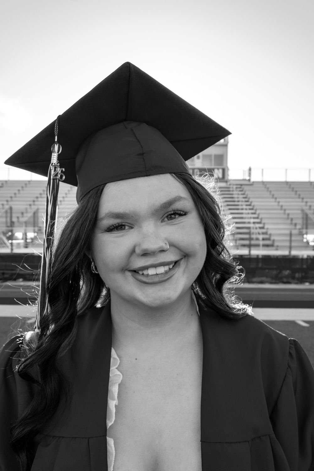 Black and white portrait of a smiling young woman in graduation cap and gown at an outdoor ceremony.