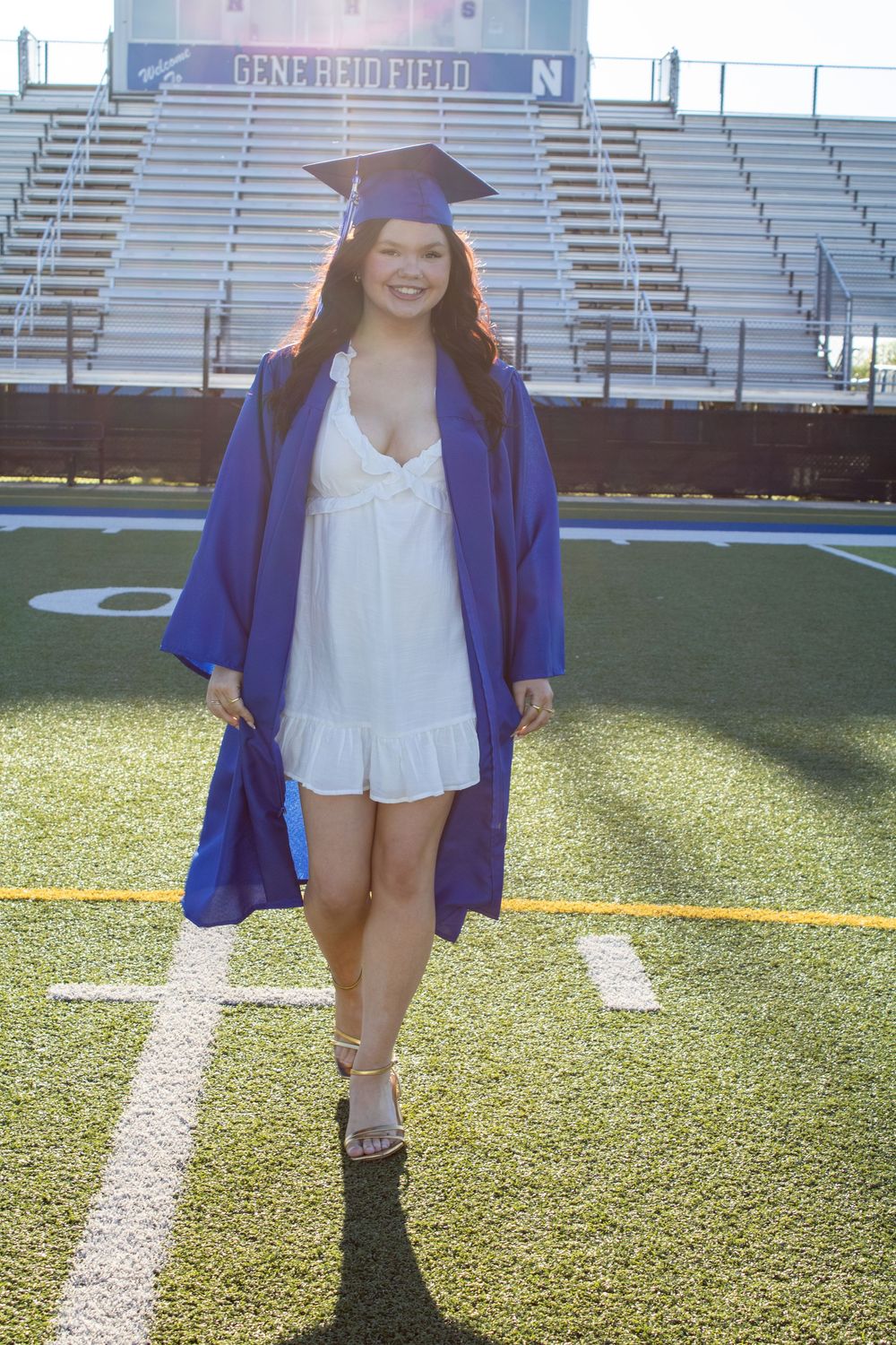 Smiling graduate in blue cap and gown over white dress poses on football field at Gene Reid Field stadium.