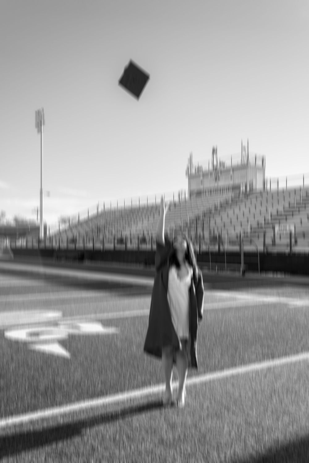 Graduate in cap and gown tosses mortarboard into air on football field, black and white photo.