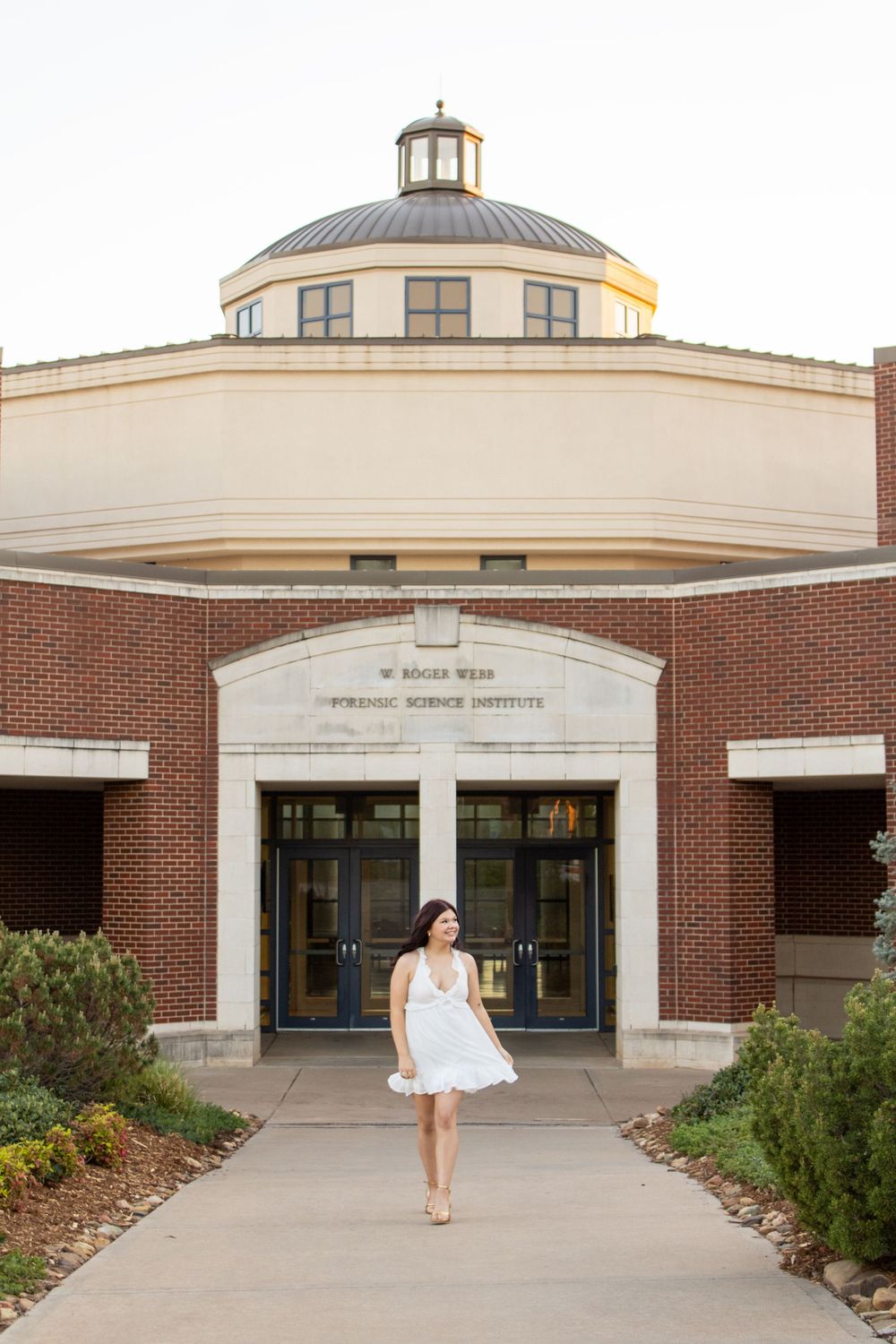 Young woman in white dress posing in front of W. Roger Webb Forensic Science Institute brick building with dome.