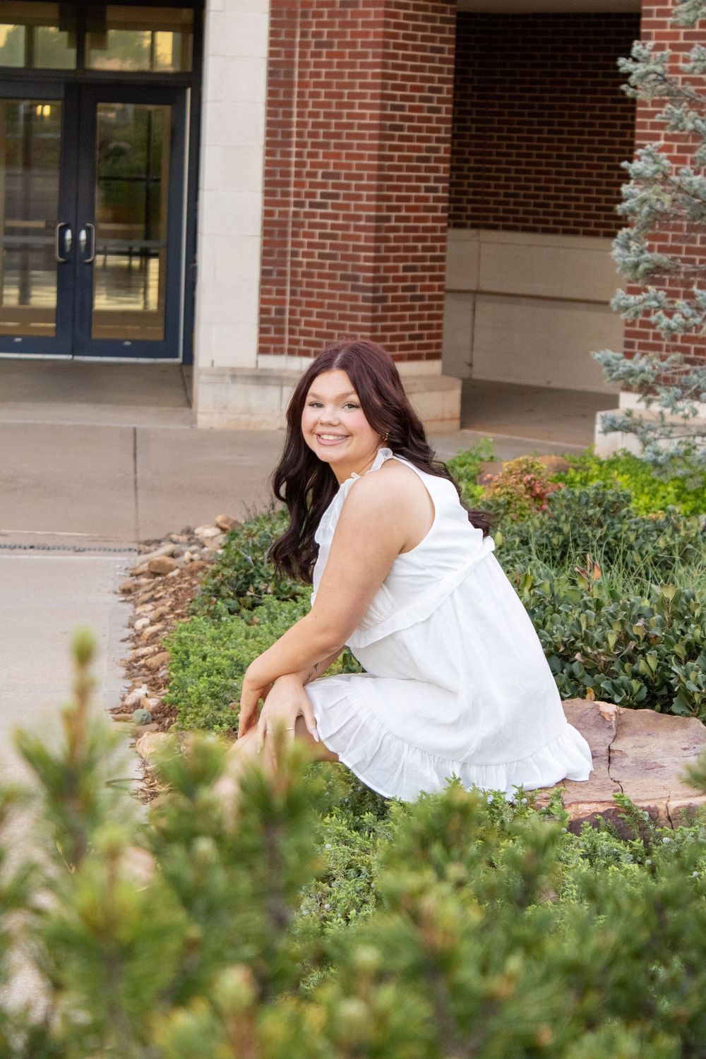 Young woman in white dress crouching by garden outside brick building, smiling over her shoulder.