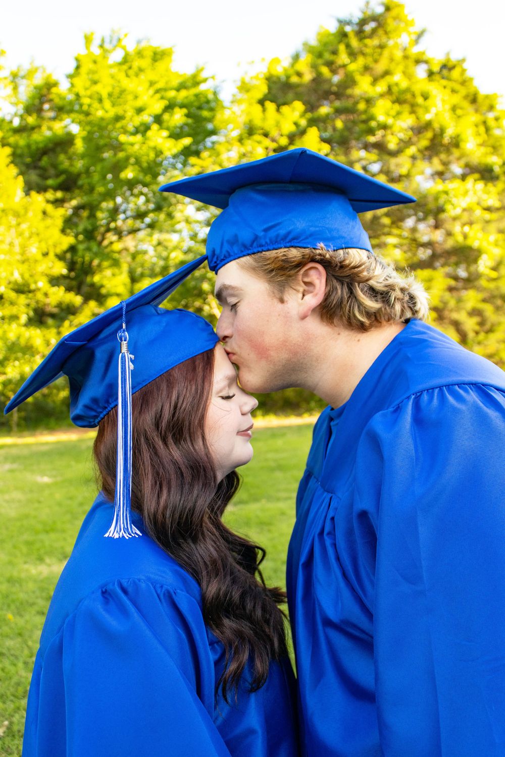Two graduates in blue cap and gown share a kiss outdoors with lush green trees in the background.