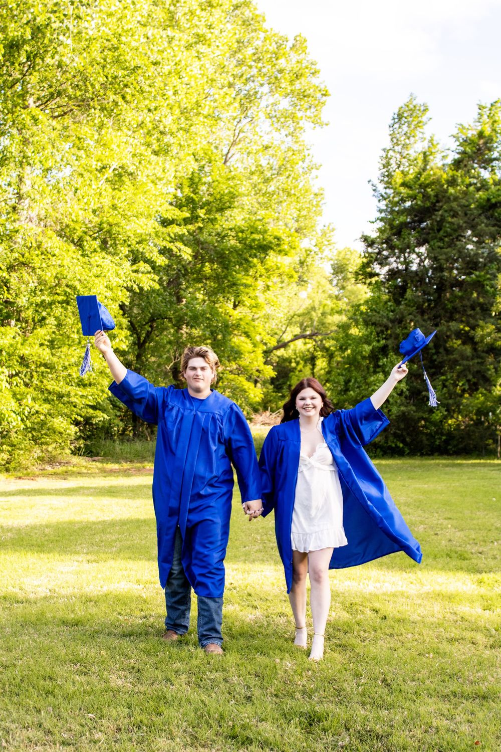 Two graduates in blue caps and gowns celebrate outdoors, holding caps high while walking hand in hand on green grass.