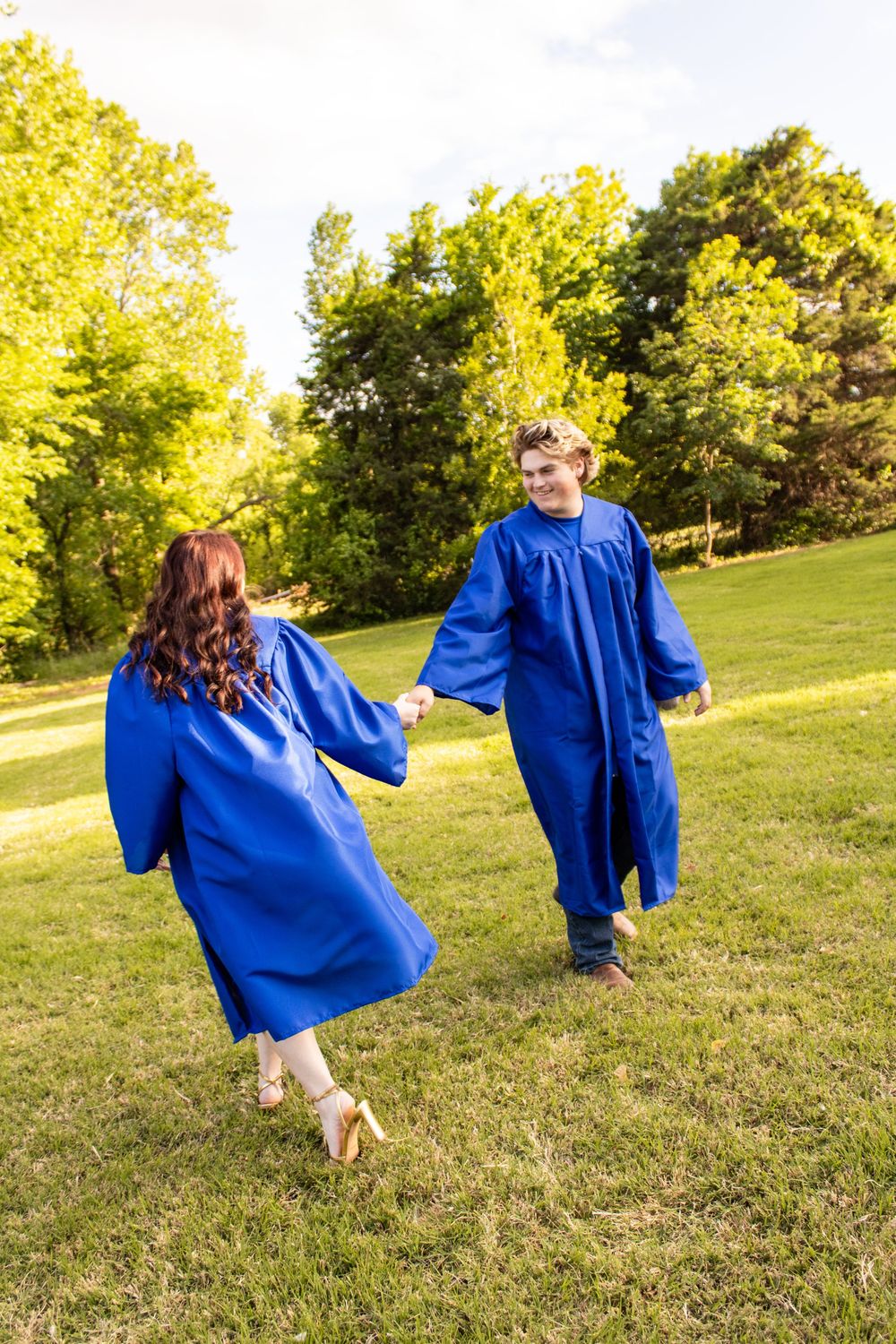 Two graduates in blue caps and gowns hold hands while walking outdoors on green grass with trees in the background.