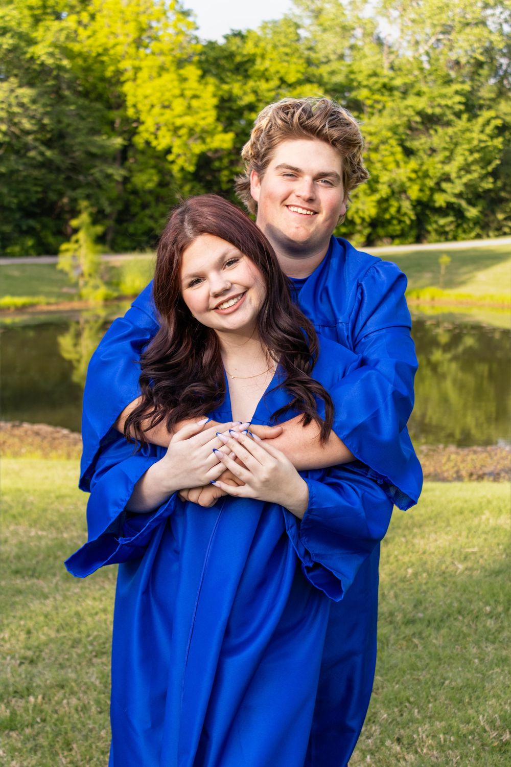 Two graduates in royal blue gowns embrace outdoors near a pond, smiling for a graduation photo.