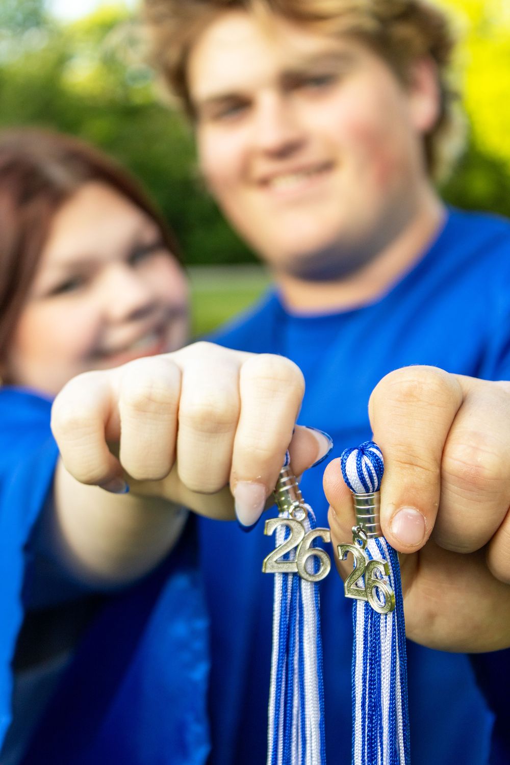 Two smiling graduates in blue shirts proudly display their 2024 tassel charms and blue and white graduation cords outdoors.