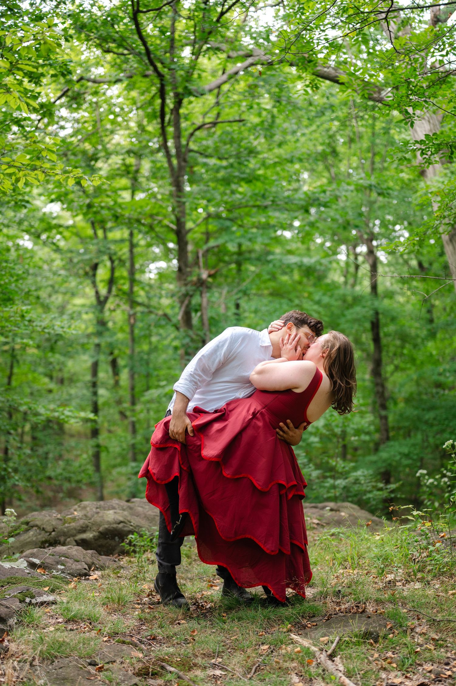Couple sharing a romantic dip kiss in a lush green forest in New Jersey, woman in flowing red gown, man in white shirt and boots.
