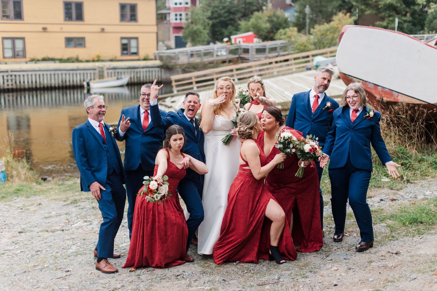 Joyful bride in a white gown surrounded by laughing groomsmen in navy suits and bridesmaids in red dresses posing by a waterfront.