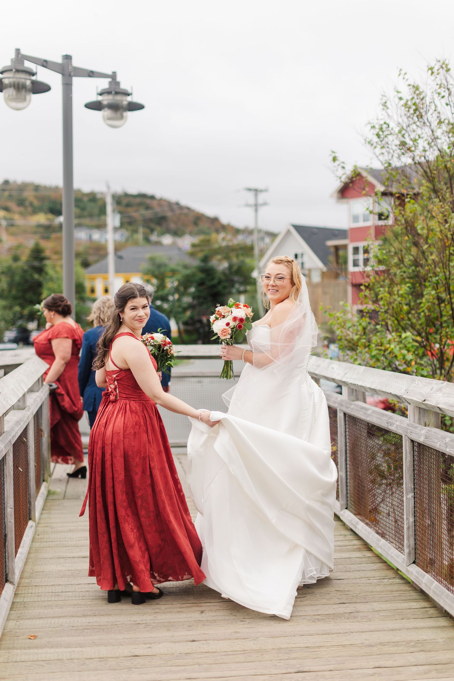 A bride in a flowing white gown holds a bouquet while a bridesmaid in a red dress holds her train on a wooden boardwalk.