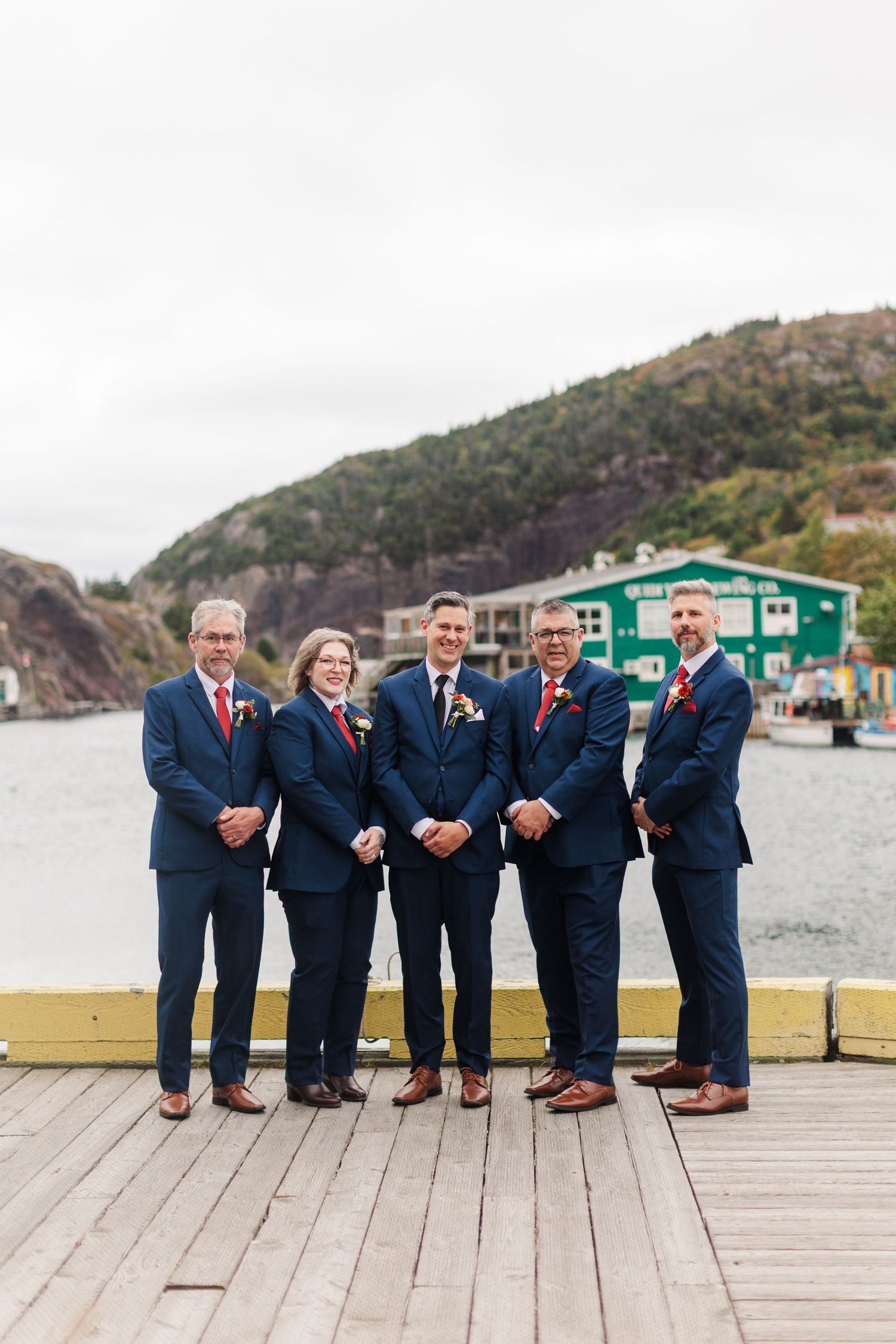 Five groomsmen in navy suits and red ties stand arm in arm on a wooden wharf with a harbor, green building and hillside behind them.