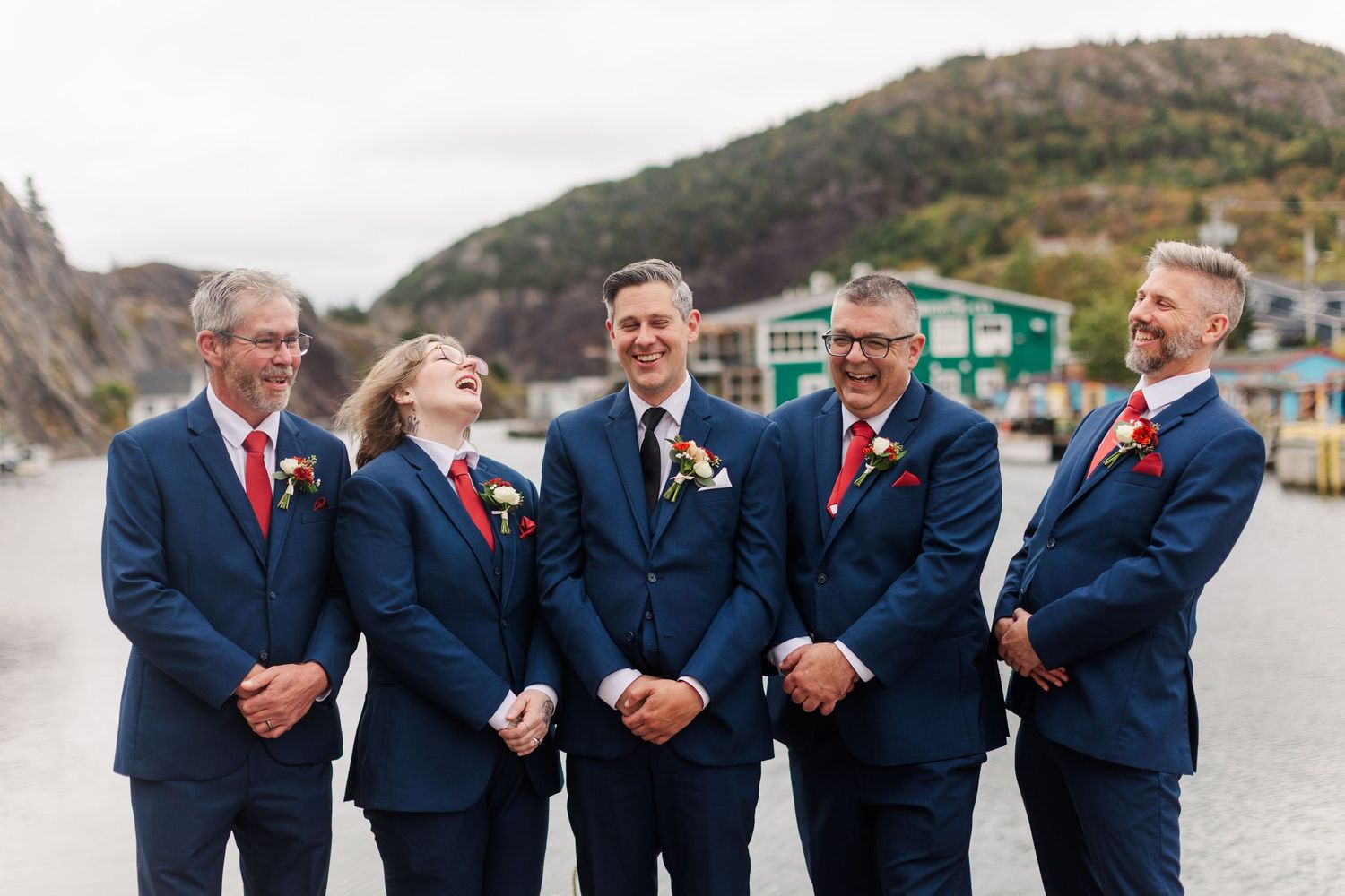 Five groomsmen in navy suits with red ties and boutonnieres laugh together on a waterfront dock with colorful buildings and hills behind.