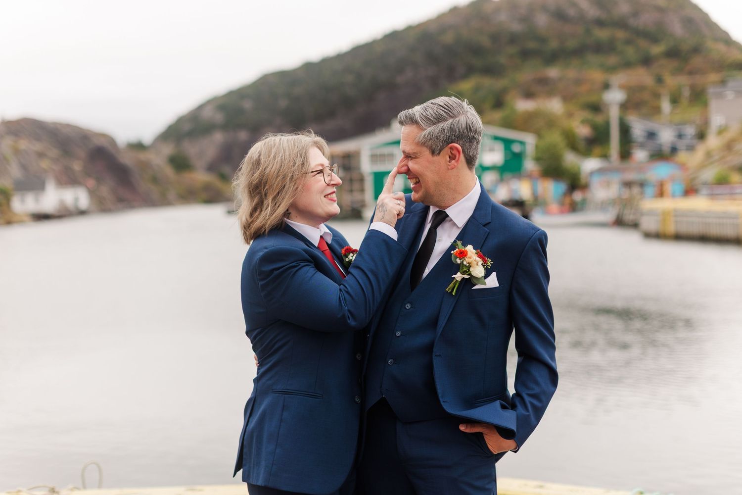 Two people in navy suits share a tender moment at a waterfront, one adjusting the other's collar, with hills and colorful buildings behind.