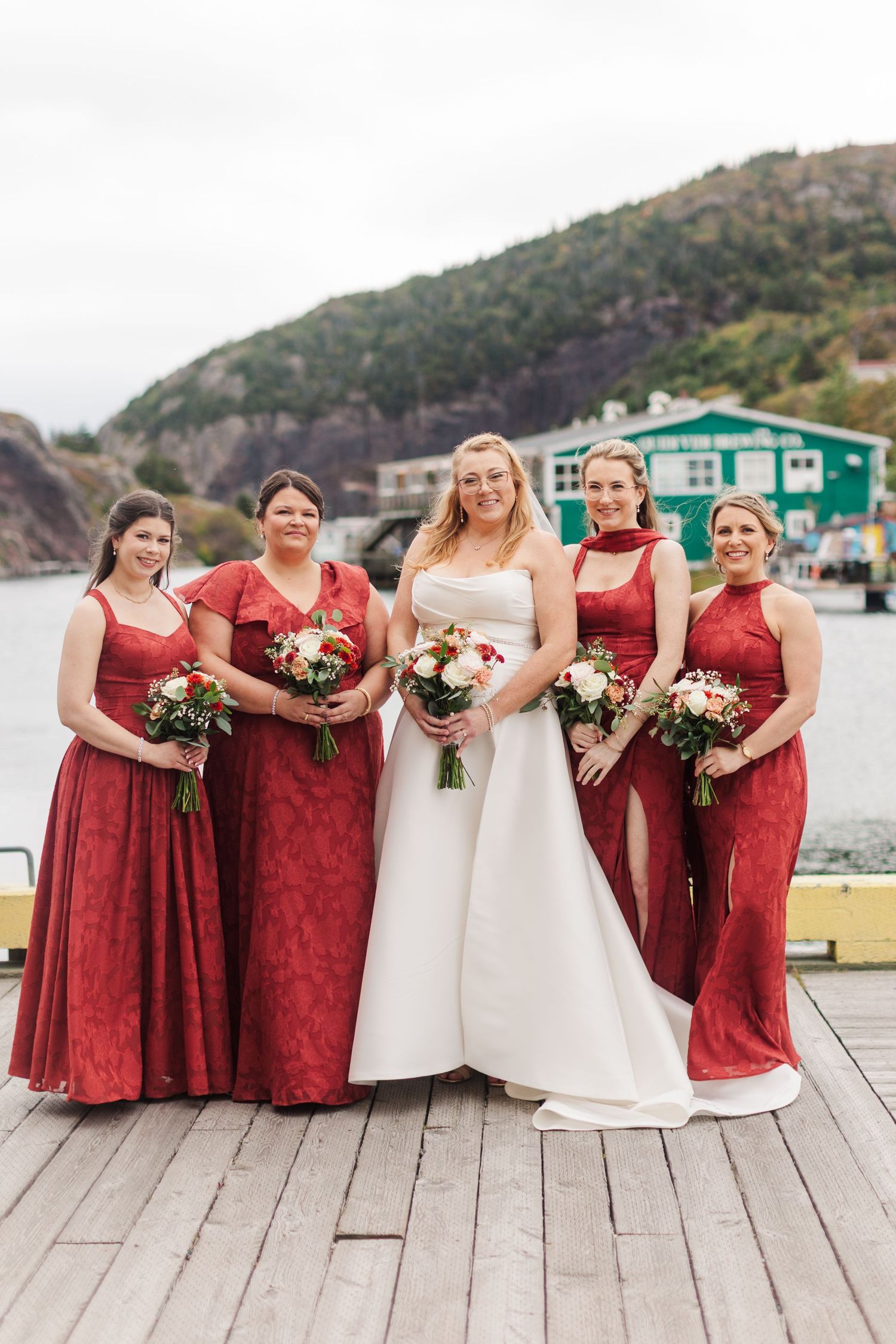 Bride in a white gown stands with four bridesmaids in red dresses holding bouquets on a wooden dock by the water.