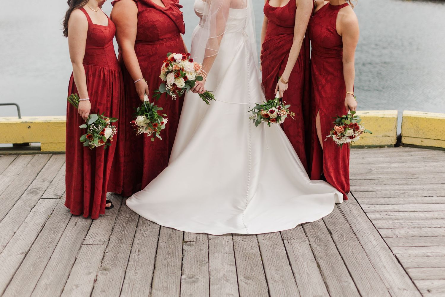 Bride in a white wedding gown stands on a wooden dock with four bridesmaids in red dresses, all holding floral bouquets by the water.