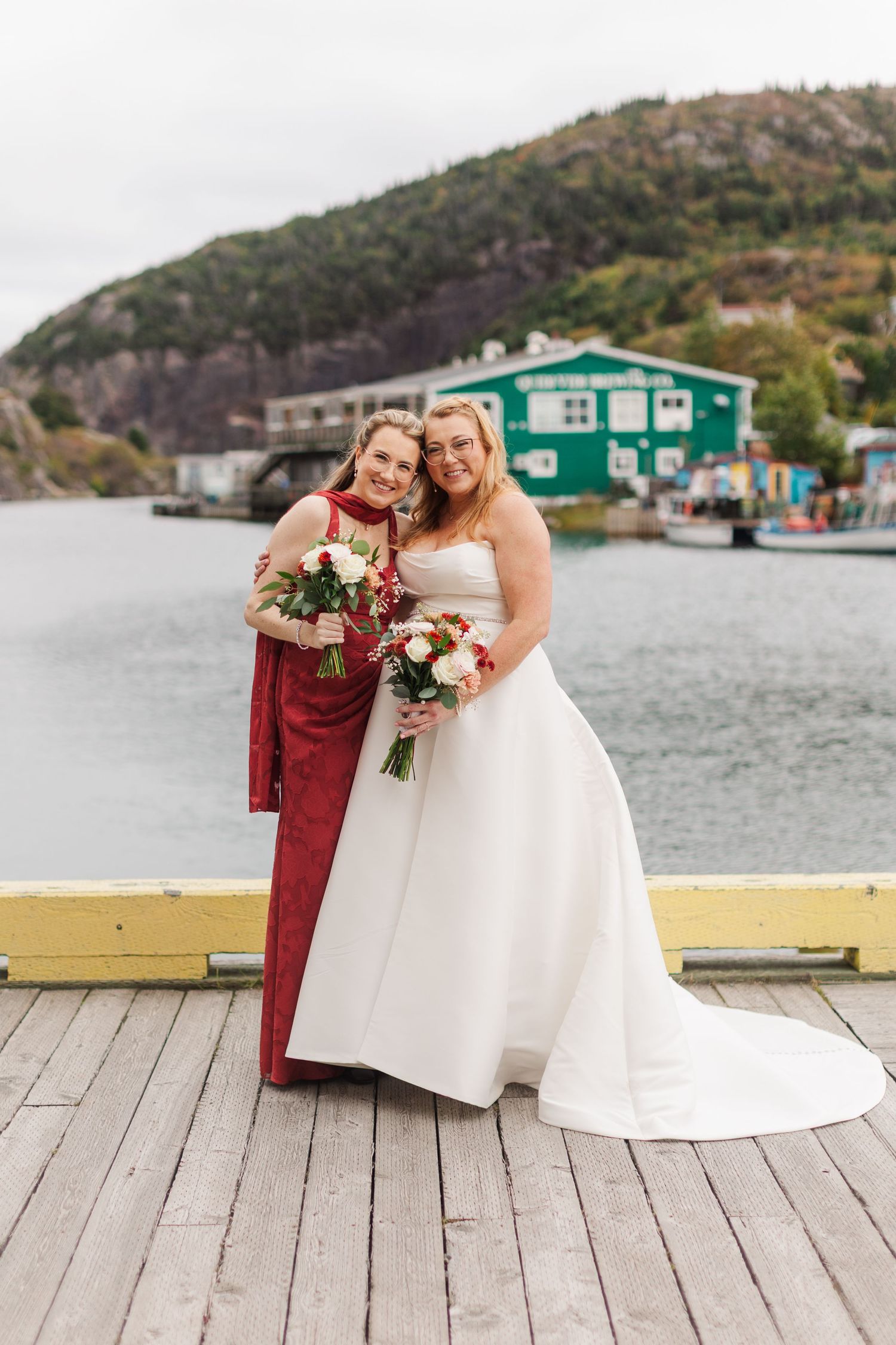 Bride in a white strapless gown and a woman in a red dress hold bouquets on a wooden waterfront dock with green buildings and hills behind.