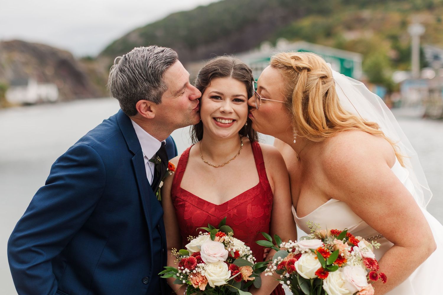 Bride in a white gown and veil and a man in a navy suit kiss the cheeks of a smiling bridesmaid in red holding a bouquet by the waterfront.