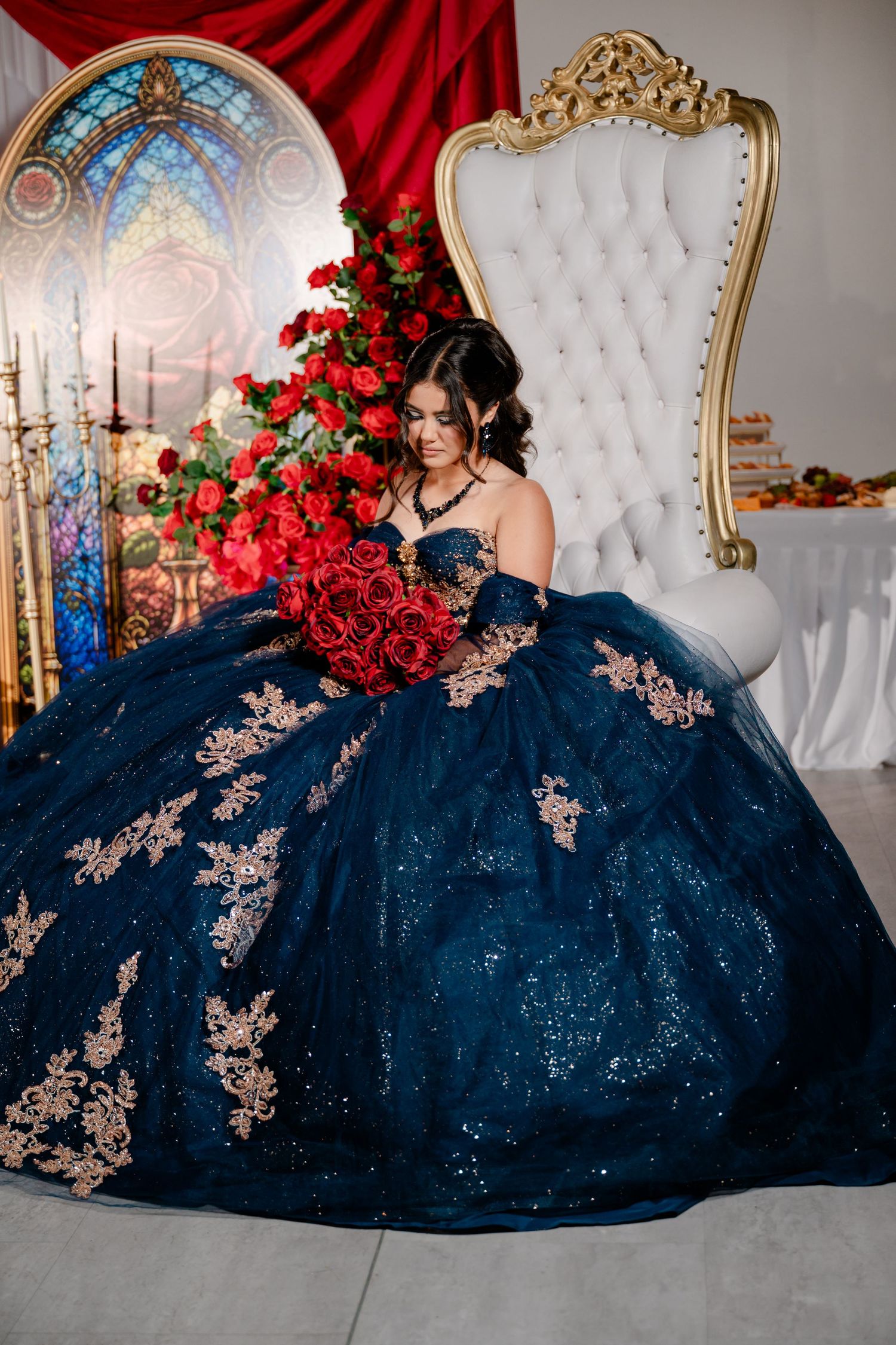 Woman in a sparkling navy blue ball gown with gold lace appliques holds a red rose bouquet while seated near a white tufted throne chair.
