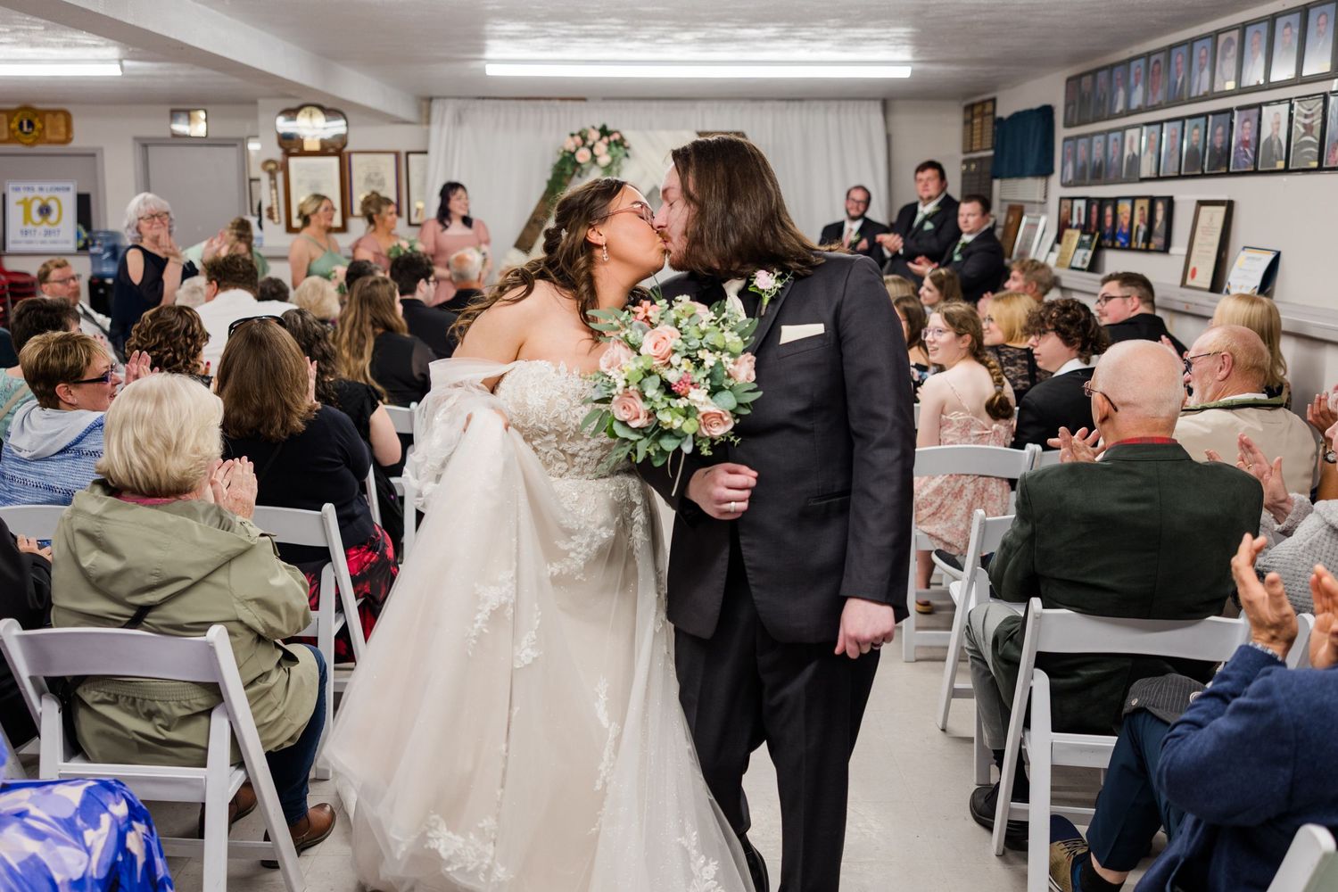 Bride and groom share a first kiss at wedding ceremony while guests applaud in decorated venue.