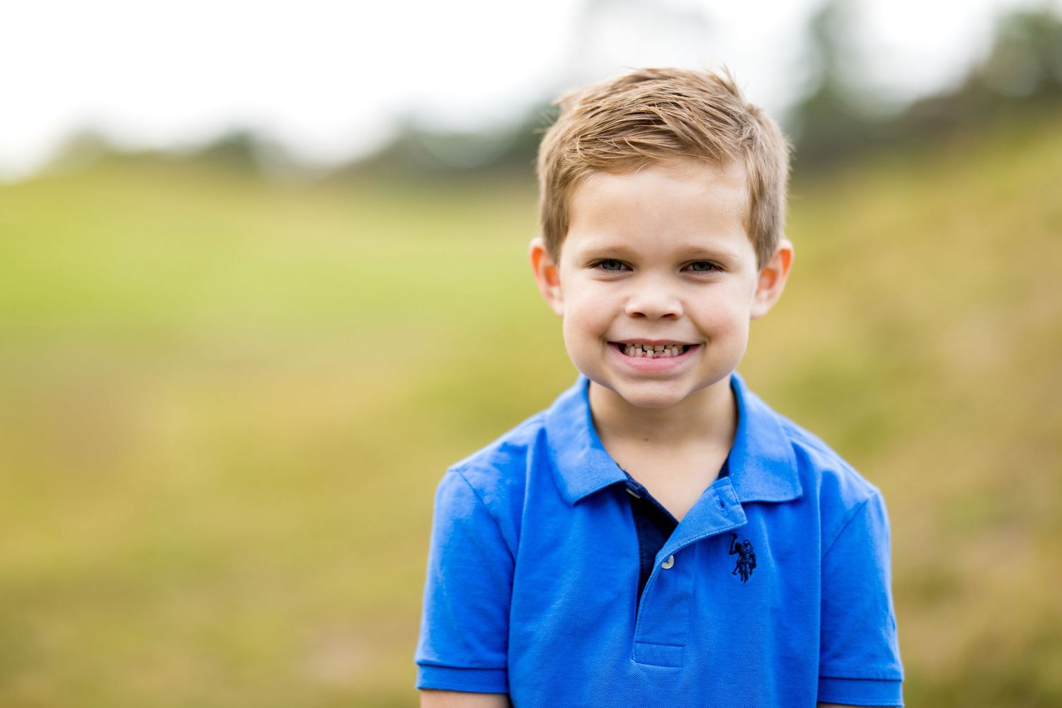 A smiling young boy in a blue polo shirt poses outdoors on a green grassy field on a sunny day.
