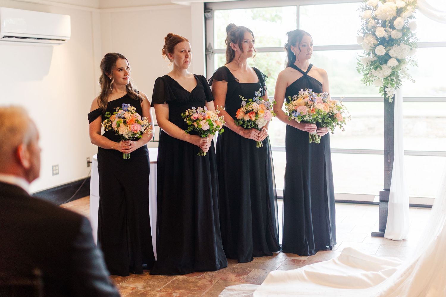 Four bridesmaids in black gowns holding colorful bouquets stand at a wedding ceremony near a floral arrangement.
