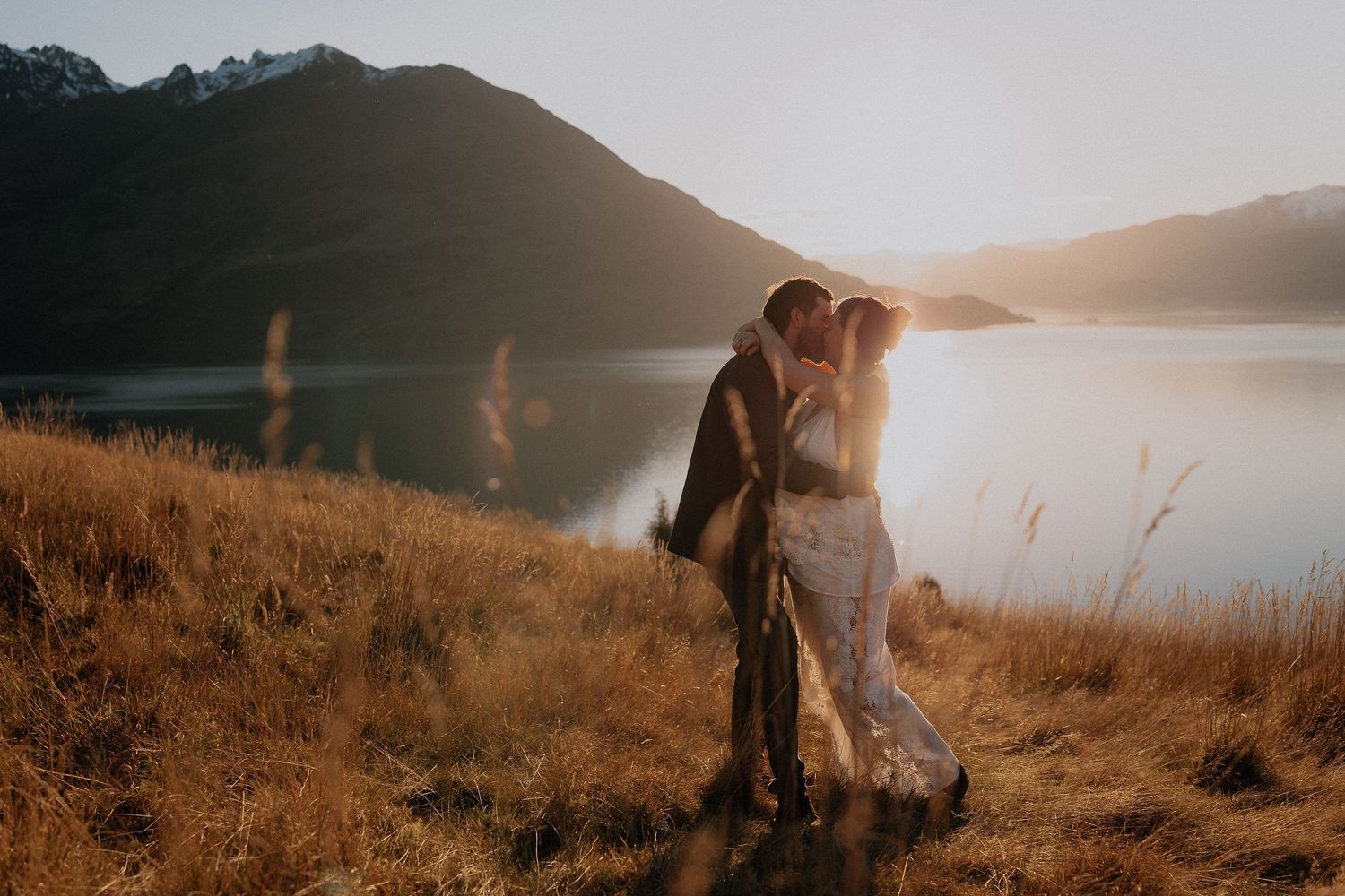 Couple sharing a romantic kiss at golden hour by a mountain lake in New Zealand with warm sunlight glowing behind them.