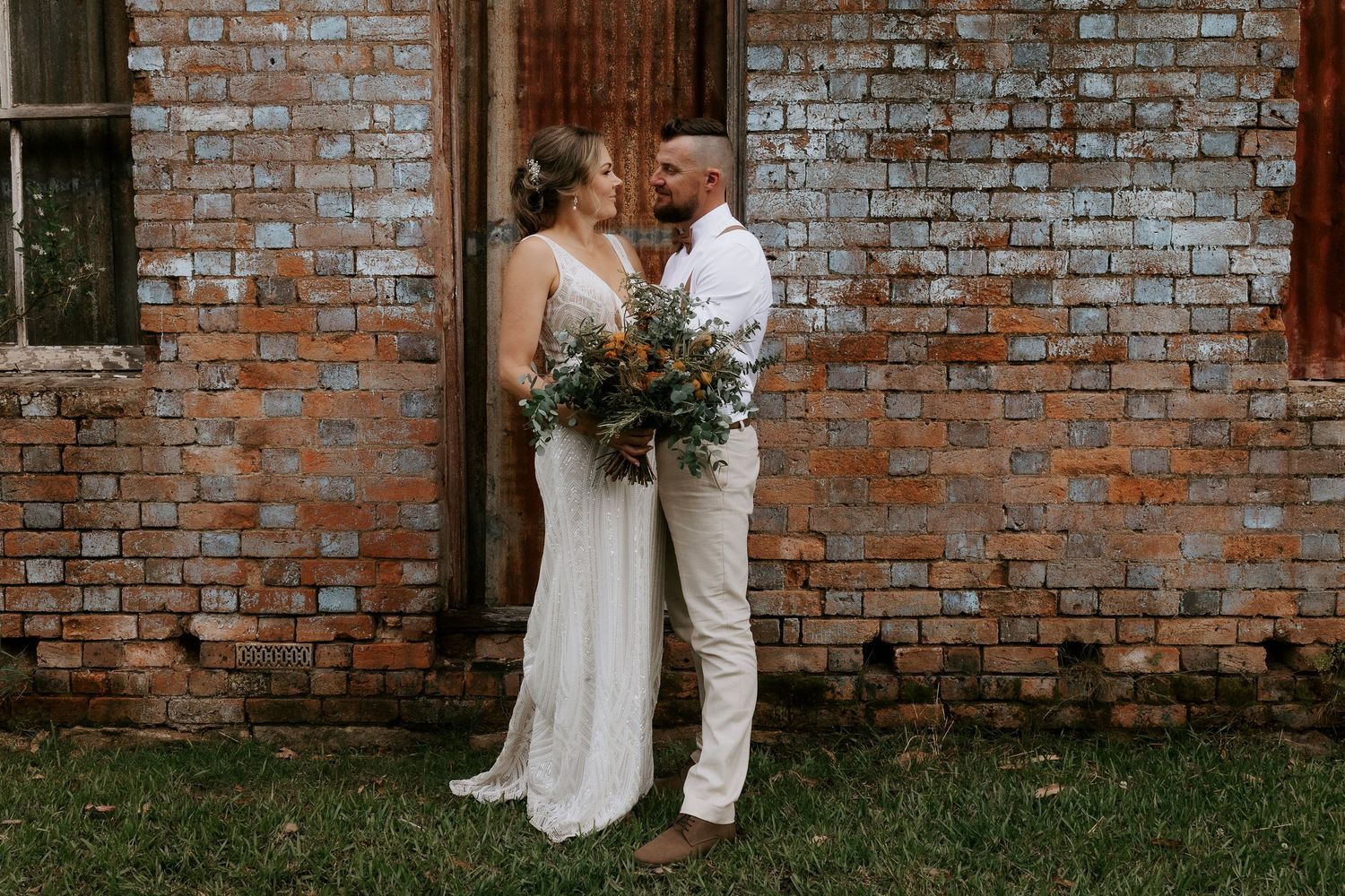Wedding couple poses against rustic brick wall in outdoor garden setting.
