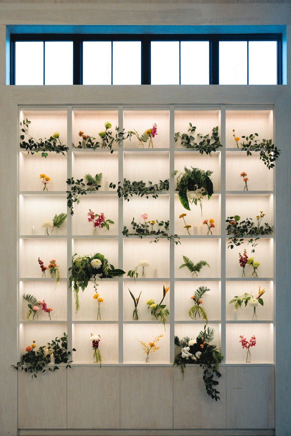 Illuminated white shelving wall displaying fresh flowers and trailing greenery in small vases beneath a high window.