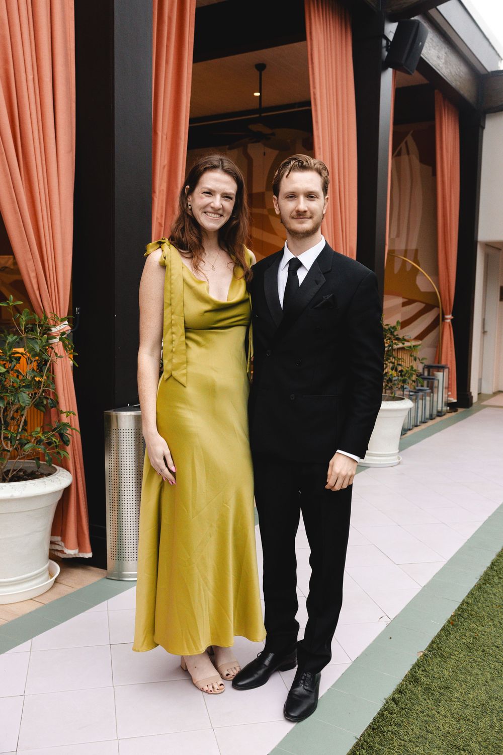 A smiling woman in a yellow satin dress poses beside a man in a black suit on a patio with coral curtains.