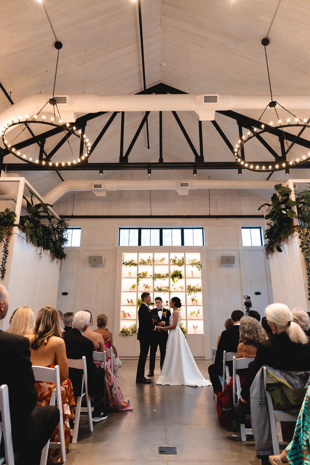Bride and groom exchange vows before seated guests in a bright modern venue with exposed wood beams, ring chandeliers and greenery.