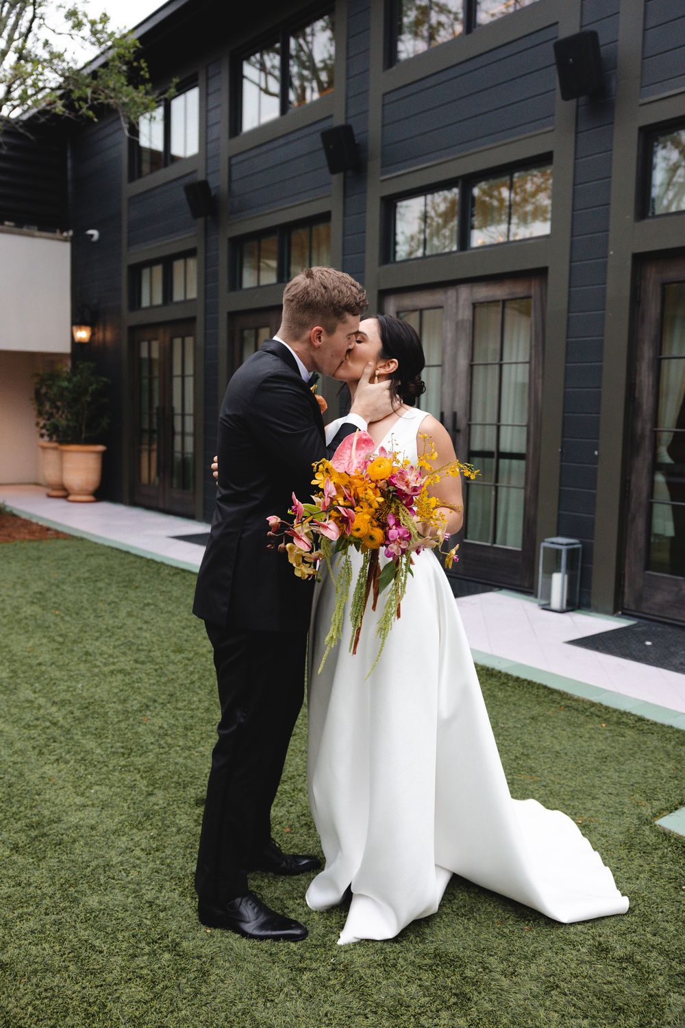 Bride in a white gown holding a colorful bouquet kisses her groom in a black suit on a green lawn outside a modern dark building.