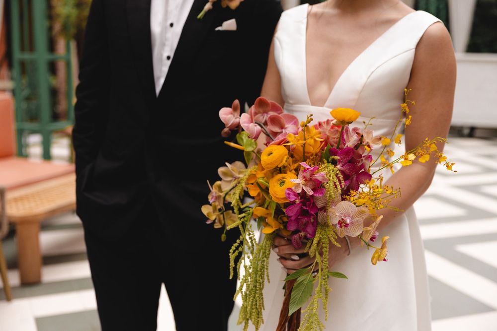 Bride in a white V-neck wedding gown holding a colorful bouquet of pink, orange and yellow flowers beside a groom in a black suit.