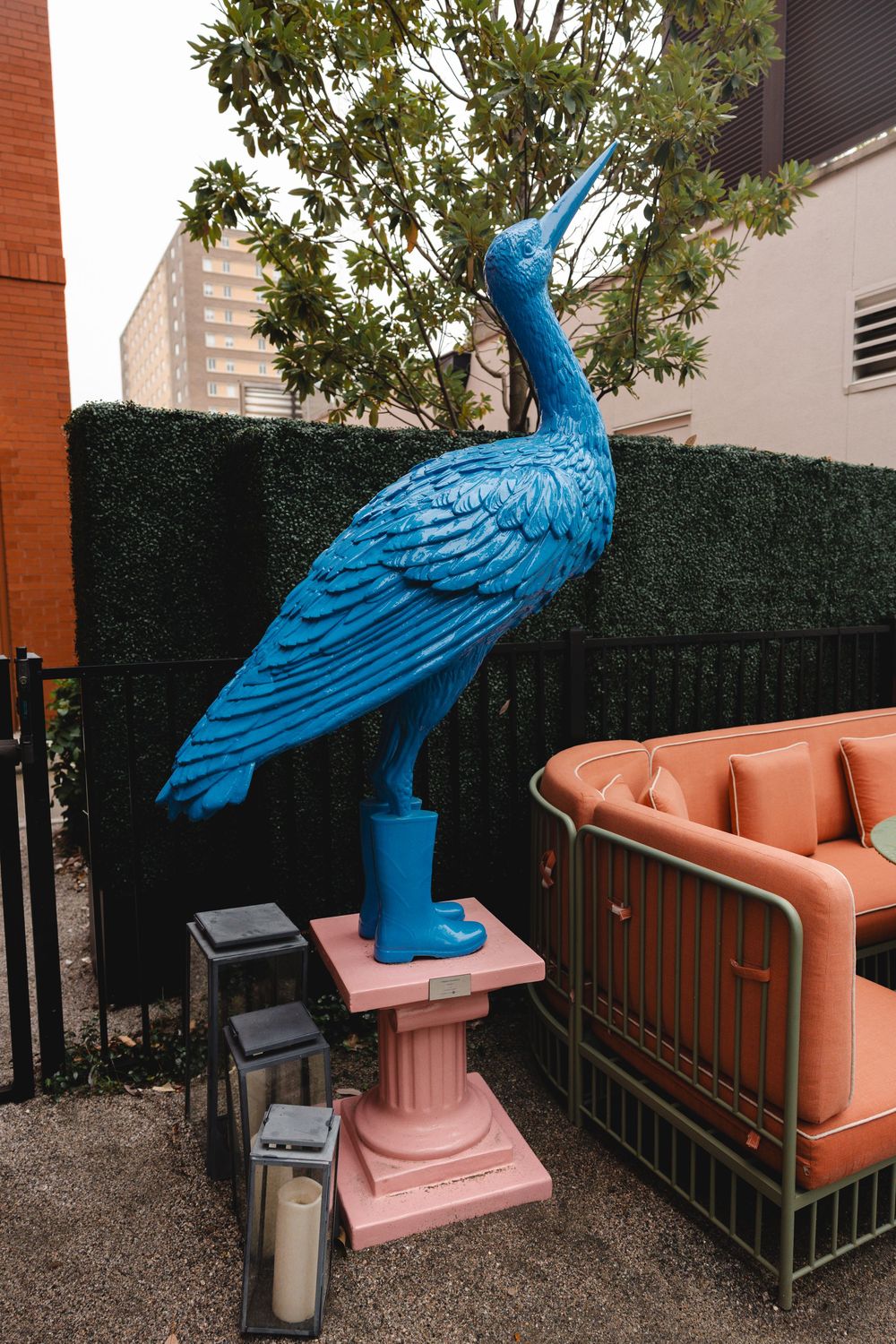 Large blue heron sculpture in blue rain boots on a pink pedestal beside an orange outdoor sofa, lanterns, and a green hedge.