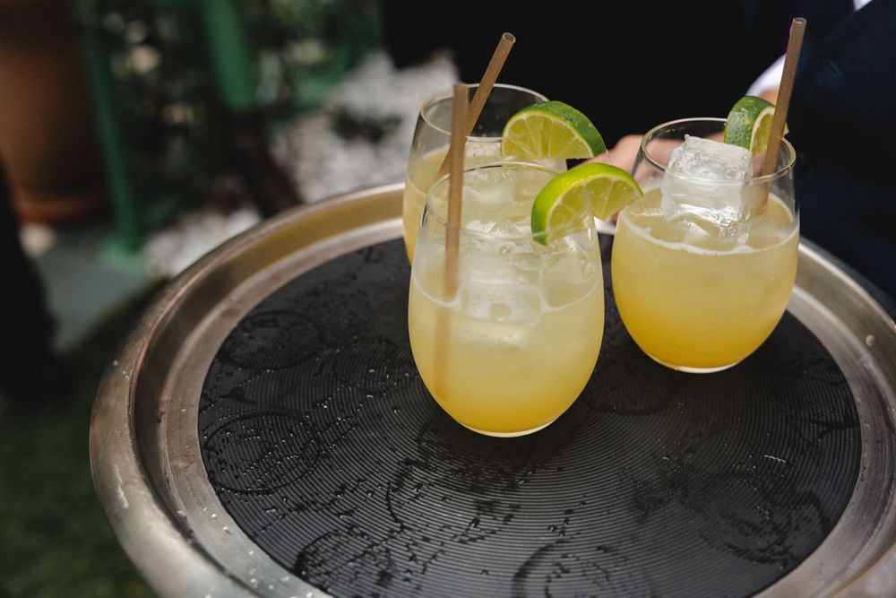 Three stemless glasses of yellow citrus cocktails with ice, lime wheels and bamboo straws on a round black serving tray.