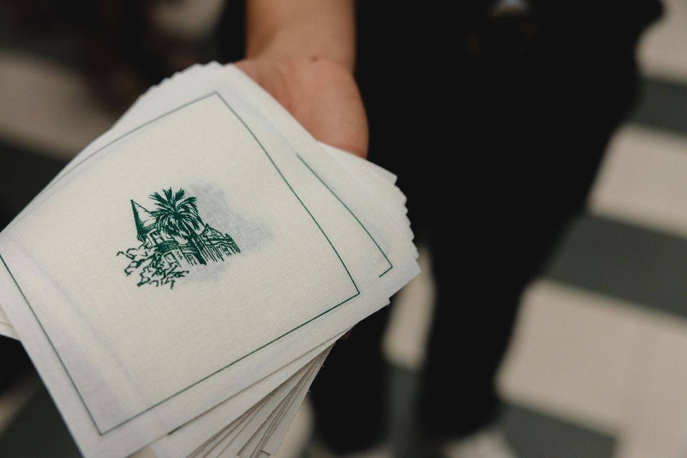 Hand holding a stack of white cocktail napkins printed with a green palm tree illustration and a green border.