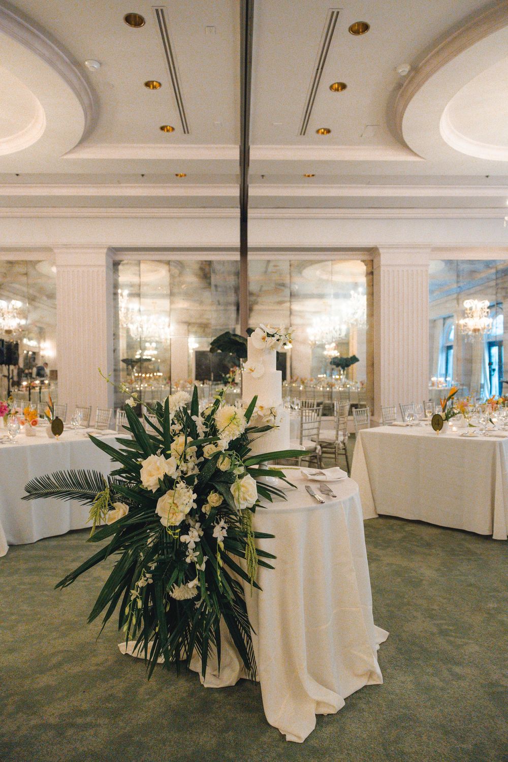 Elegant ballroom wedding reception with a floral centerpiece of white blooms and palm leaves on a round table, set beneath chandeliers.