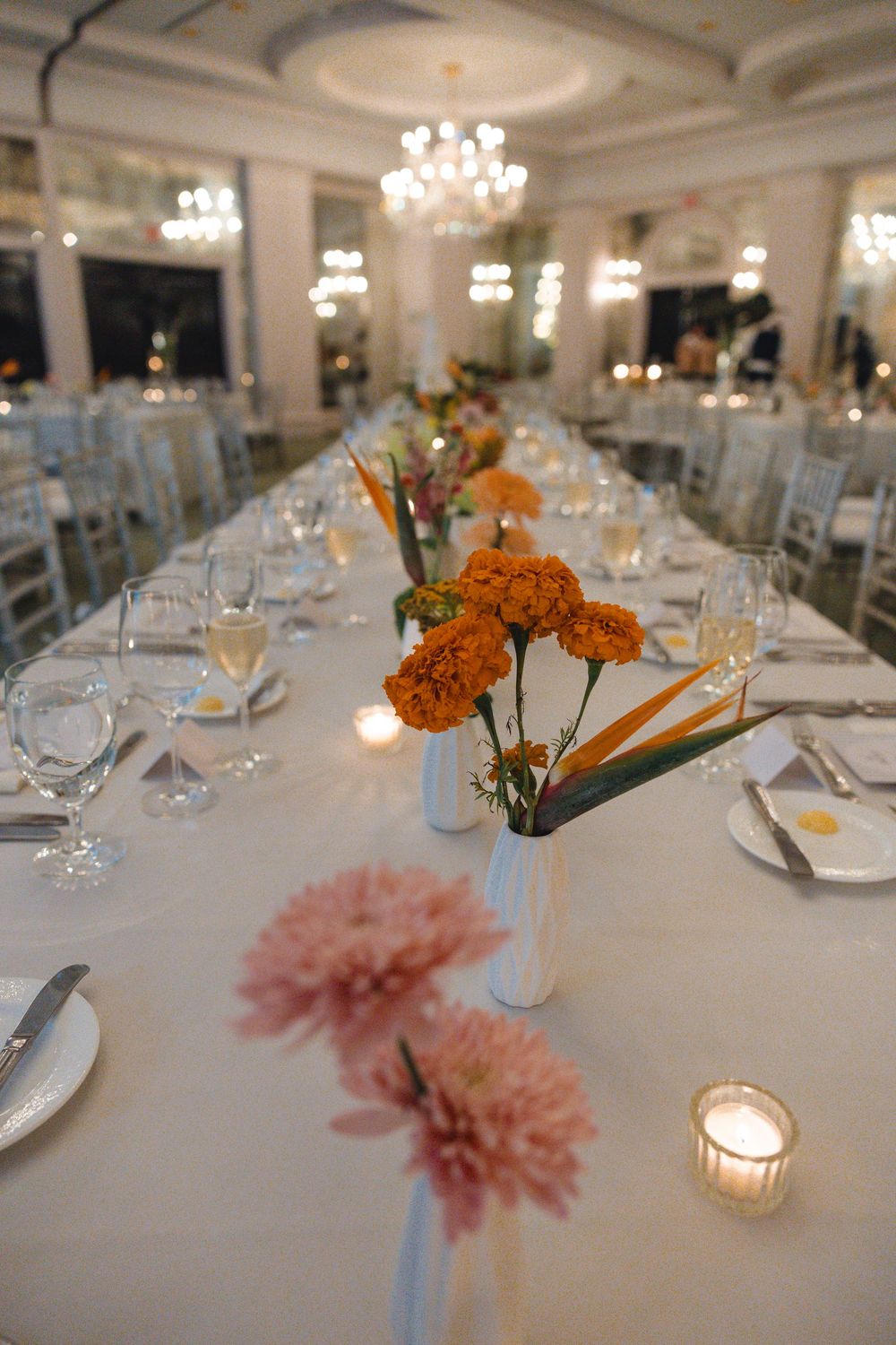 Elegant banquet table with white linens, orange marigold and pink bloom centerpieces, candles and glassware beneath a crystal chandelier.