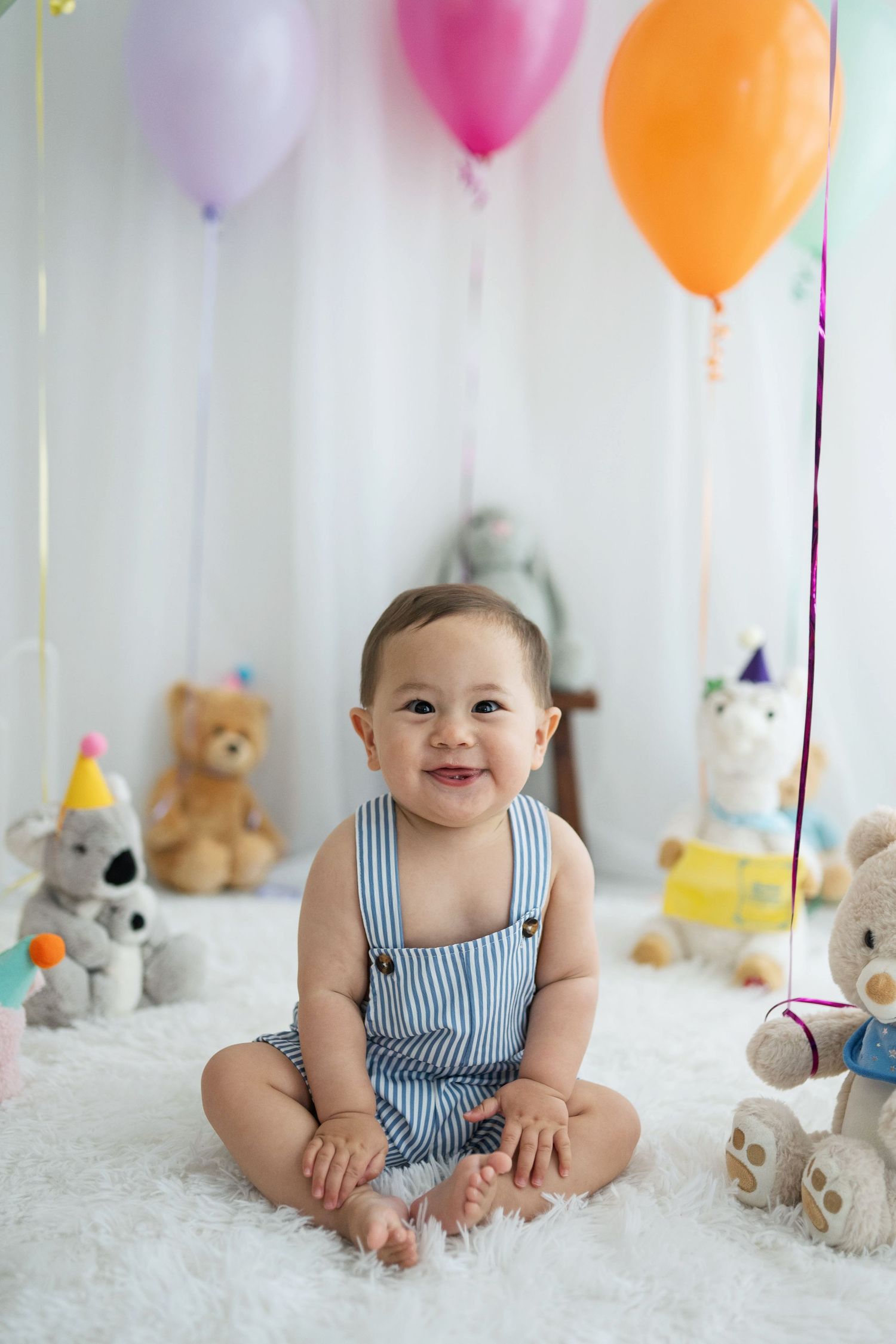 Smiling baby in striped overalls sits on white fur rug surrounded by stuffed animals and colorful balloons at birthday party.